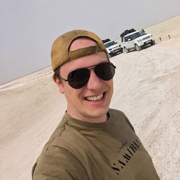 Sebastian mit Cap und Sonnenbrille auf einer Wüstenpiste, dahinter zwei Geländewagen